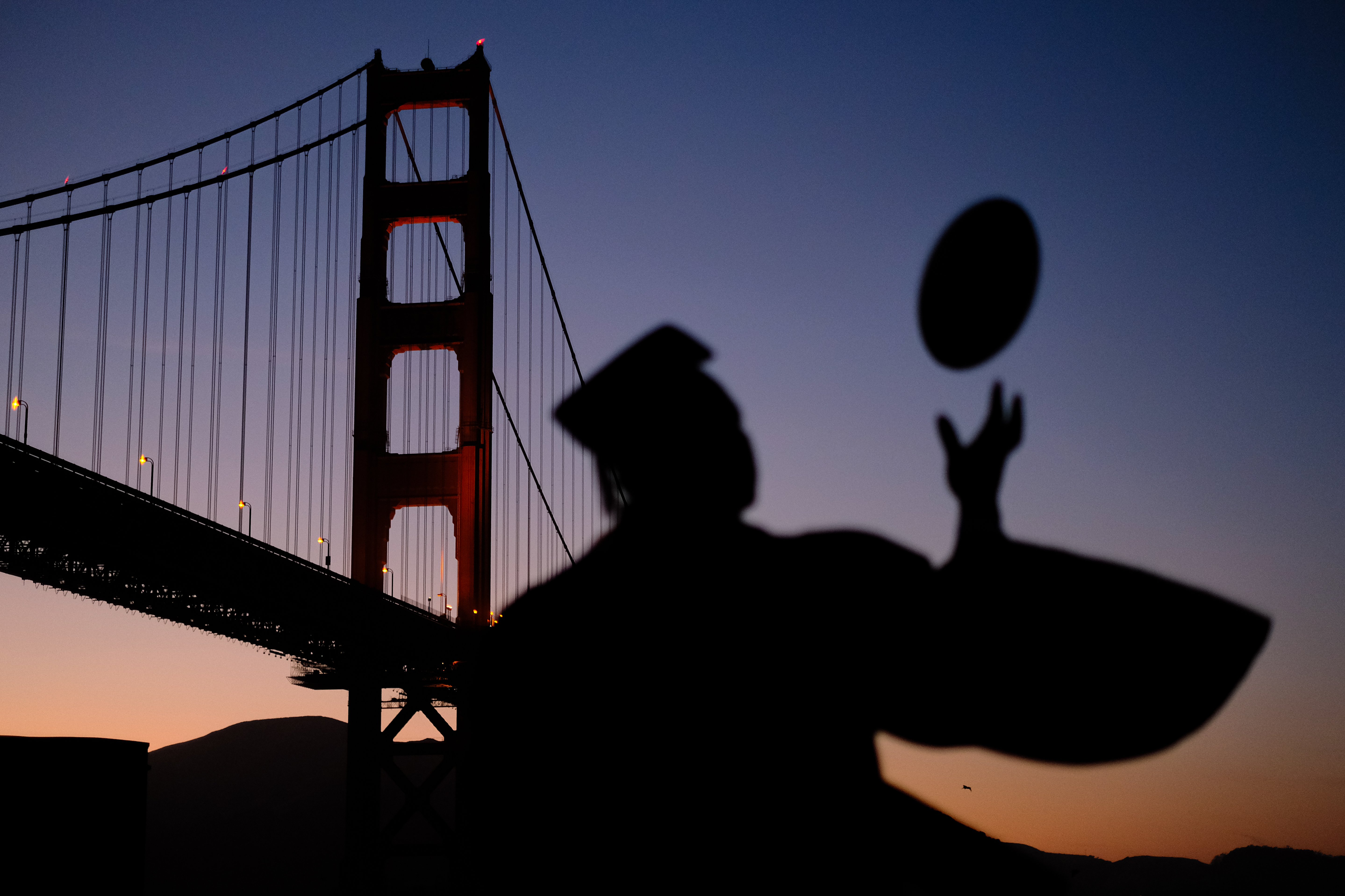 Student with graduation cap on stands in front of the Golden Gate Bridge while tossing a graduation cap in the air. This is at dawn, it is starting to get dark out